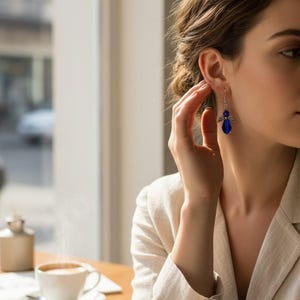 May include: A woman wearing a light-colored blazer and blue earrings. The earrings are a dark blue color and have a decorative design. A cup of coffee sits on a table in the foreground.