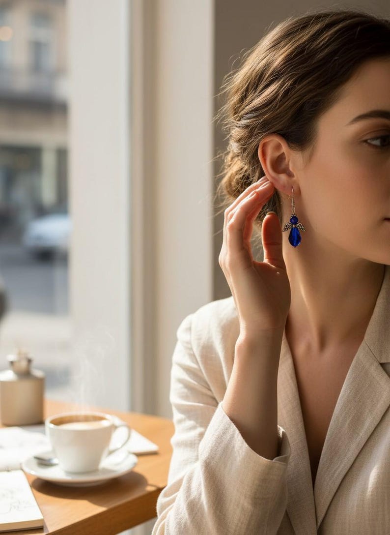 May include: A woman wearing a light-colored blazer and blue earrings. The earrings are a dark blue color and have a decorative design. A cup of coffee sits on a table in the foreground.