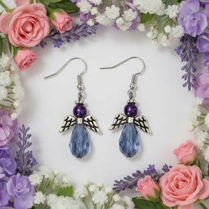 May include: A pair of silver-toned angel earrings with purple beads and blue teardrop-shaped crystals. The earrings have detailed wing designs and are set against a backdrop of pink and purple flowers.