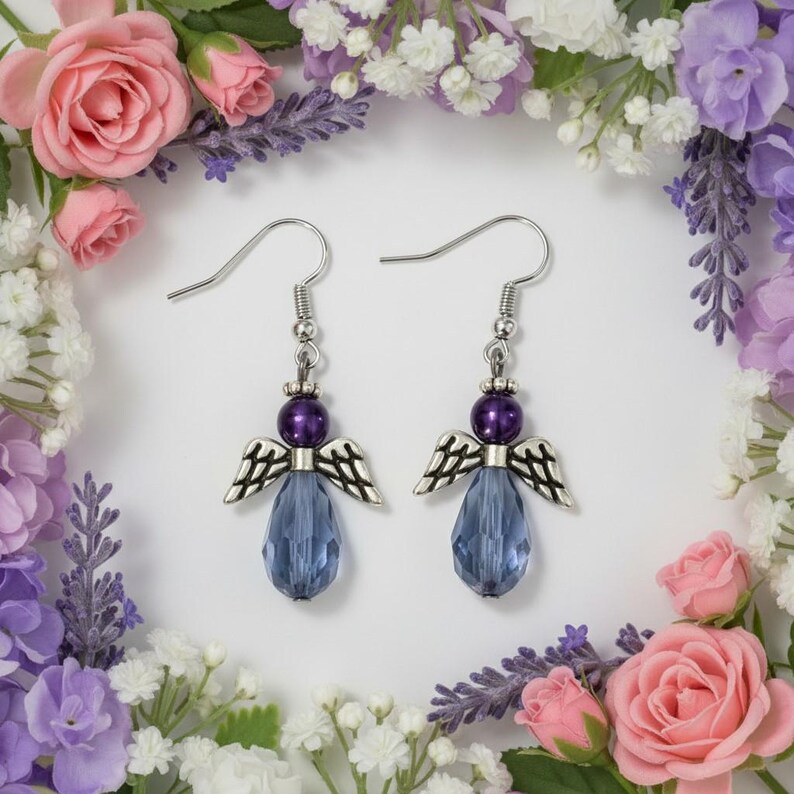 May include: A pair of silver-toned angel earrings with purple beads and blue teardrop-shaped crystals. The earrings have detailed wing designs and are set against a backdrop of pink and purple flowers.