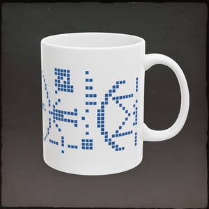 May include: White ceramic mug with a blue pixelated design. The design appears to be an abstract pattern of squares and lines. The mug has a curved handle and is set against a dark background.