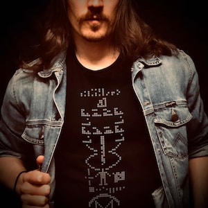 May include: Black t-shirt with a pixelated design and a denim jacket. The t-shirt features a white, pixelated graphic. The denim jacket is light blue and has a button closure. The image is set against a dark background.