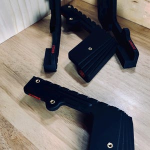 Puede incluir: Soportes de montaje negros con detalles rojos y las palabras "SINGLE" y "DOUBLE" impresas. Los soportes están hechos de un material acanalado y tienen herrajes dorados. Los soportes se muestran sobre una superficie de madera.