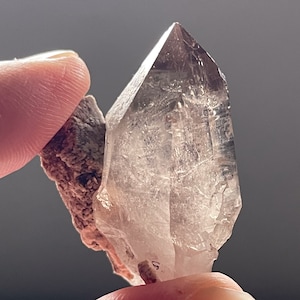 Puede incluir: Un cristal de cuarzo transparente con una punta puntiaguda, unido a una roca marrón y gris. El cristal es translúcido y tiene un ligero brillo.