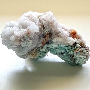 May include: A natural mineral specimen with a light blue-grey, crystalline structure. The mineral has areas of brown and orange hues, with a turquoise-colored base. The overall shape is irregular and textured, showcasing natural geological formations.