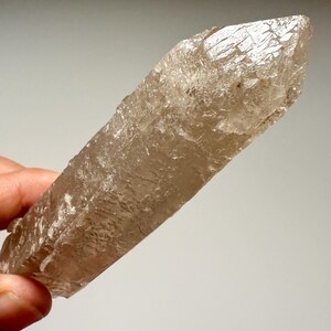May include: A translucent, light brown crystal with a textured surface. The crystal is elongated with a pointed tip. The overall shape is rectangular, with a rough, natural appearance. The crystal is held in a hand.