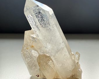 May include: A clear quartz crystal cluster with multiple points. The crystal is transparent with subtle internal fractures and a light, natural color. The cluster has a rough base and a polished, pointed top.