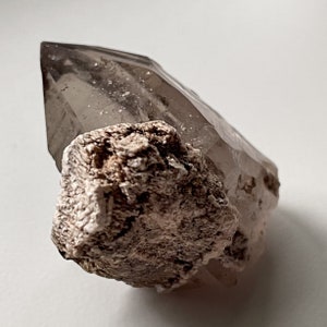 May include: A clear quartz crystal with a brown, rough, and textured base. The crystal is pointed and has a natural, raw look.