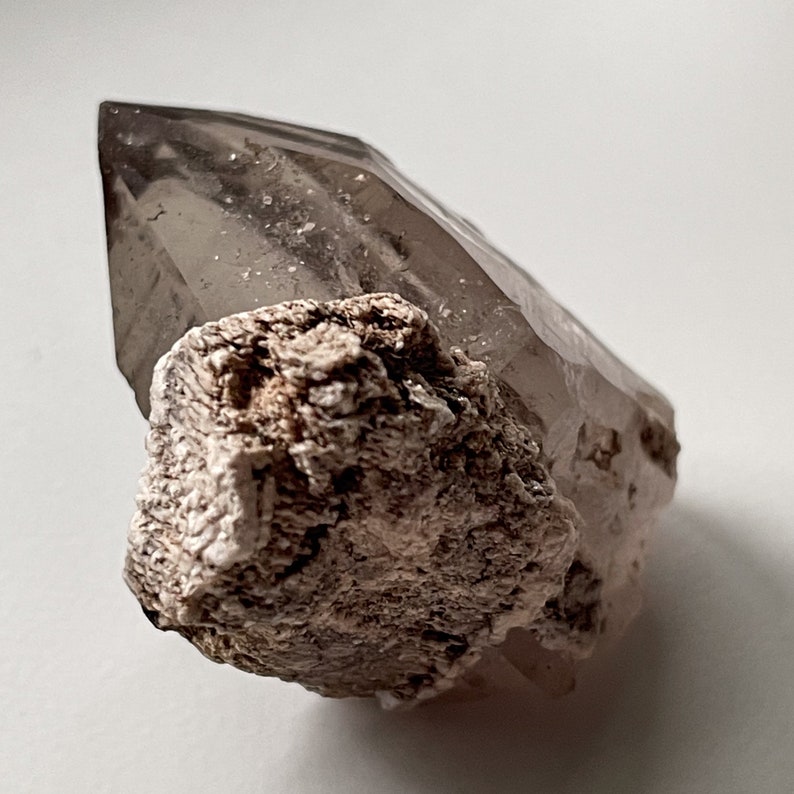 May include: A clear quartz crystal with a brown, rough, and textured base. The crystal is pointed and has a natural, raw look.