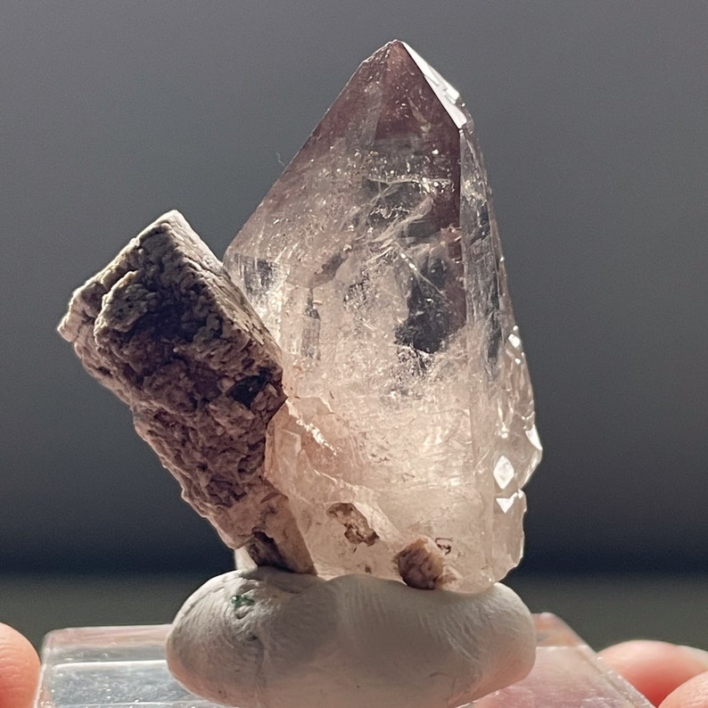 May include: A clear quartz crystal with a pink hue, mounted on a small white base. The crystal is attached to a piece of brown rock.