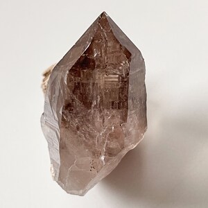 May include: A large, clear, smoky quartz crystal point with a natural, rough base. The crystal is translucent and has a light brown color.