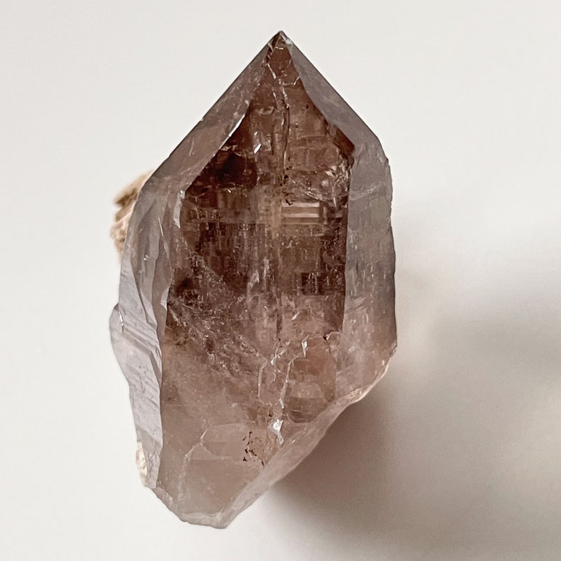 May include: A large, clear, smoky quartz crystal point with a natural, rough base. The crystal is translucent and has a light brown color.