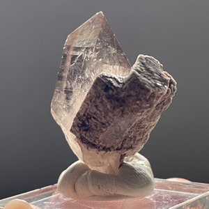 May include: A clear quartz crystal with a smoky brown hue, mounted on a white base. The crystal is shaped like a pyramid and has a sharp point. The base is made of a soft, white material.