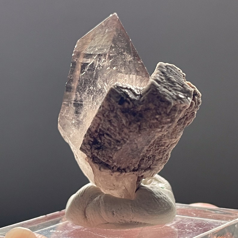 May include: A clear quartz crystal with a smoky brown hue, mounted on a white base. The crystal is shaped like a pyramid and has a sharp point. The base is made of a soft, white material.