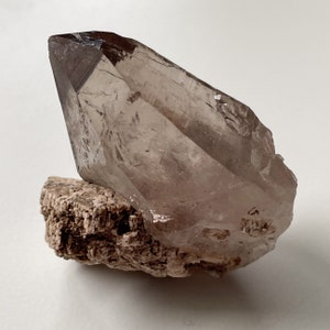 May include: A large, clear quartz crystal with a smoky brown hue, resting on a rough, brown, natural rock base.