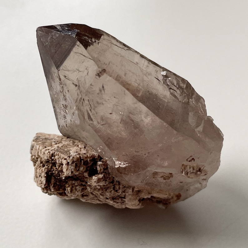 May include: A large, clear quartz crystal with a smoky brown hue, resting on a rough, brown, natural rock base.