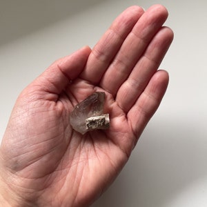 May include: A smoky quartz crystal with a natural, rough texture. The crystal is a light brown color and has a pointed shape. It is sitting in the palm of a hand.