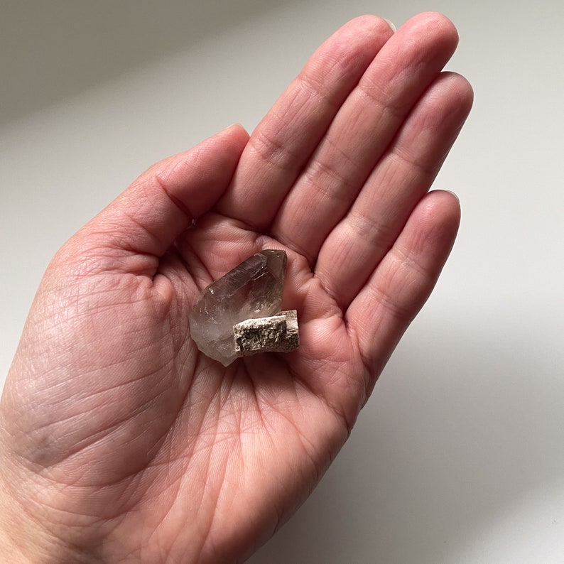 May include: A smoky quartz crystal with a natural, rough texture. The crystal is a light brown color and has a pointed shape. It is sitting in the palm of a hand.