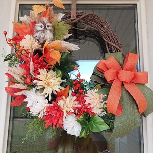 May include: A fall-themed wreath with a grapevine base, adorned with orange, red, and white flowers, fall leaves, and berries. A small owl figurine is nestled among the foliage. An orange bow with green backing completes the design.