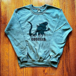 Può includere: Una felpa blu con una silhouette nera di Godzilla che distrugge una città. Il testo "GODZILLA" è stampato sotto l'immagine.