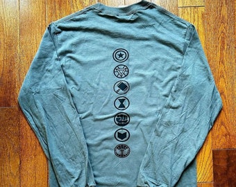 May include: A long-sleeved, teal blue t-shirt with a vertical row of seven black superhero logos printed on the back. The logos include Captain America, Iron Man, Thor, Black Widow, Hulk, Hawkeye, and Spider-Man.