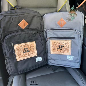 May include: Two backpacks, one black and one gray, with leather patches. The black backpack has "JL" initials, and the gray one has "JF". Both have a brown diamond-shaped patch. The backpacks are on a car seat.