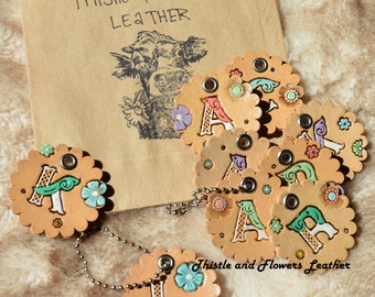 May include: A set of leather keychains with painted letters, flowers, and birds. The keychains are attached to a silver chain. The keychains are labeled "Thistle and Flowers Leather".