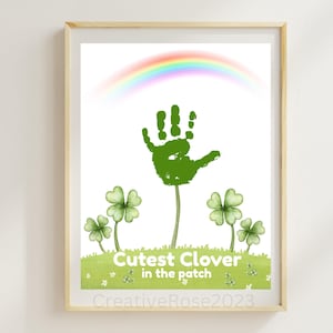 Pode incluir: Impressão emoldurada com uma impressão de mão verde em forma de trevo, com um arco-íris acima. O texto "Cutest Clover in the patch" é exibido em uma área gramada verde com trevos. A obra é em estilo aquarela.