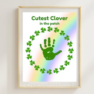 Op de afbeelding: Een ingelijste print met de tekst "Cutest Clover in the patch" in het groen. De print toont een groene handafdruk in het midden, omringd door een cirkel van groene klavers. Een regenboogverloop op de achtergrond voegt kleur toe aan het ontwerp.
