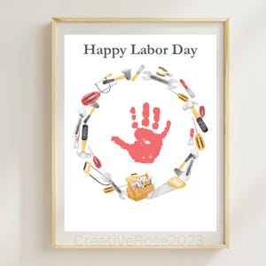 May include: A framed print with the text "Happy Labor Day." The artwork features a red handprint in the center, surrounded by a circular arrangement of tools. A small toolbox and saw are at the bottom. The frame is a light wood color.