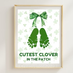 Op de afbeelding: Een ingelijste print met groene babyvoetjes, een groene strik en klavers op een witte achtergrond. De tekst "Cutest Clover in the Patch" is groen. De print zit in een licht houten frame.