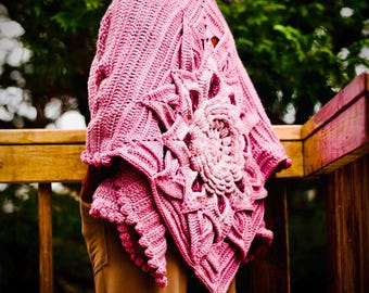 May include: A pink crocheted shawl with a large floral design on the back. The shawl has a textured pattern and a scalloped edge. The person is wearing brown pants. The background is a wooden deck and green foliage.