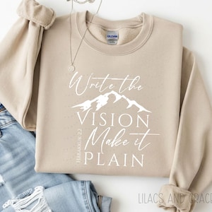 May include: A beige sweatshirt with the text "Write the Vision, Make it Plain" in white script, with a mountain graphic. The text "Habakkuk 2:2" is printed vertically on the left side. The brand name "Lilacs and Grace Co." is printed at the bottom.