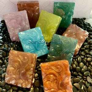 May include: Assortment of handmade soap bars with swirling patterns in various colors, including brown, pink, green, blue, and yellow. The soap bars are arranged on a bed of black pebbles. The text "Sandy's Suds" is visible in the center of the image.