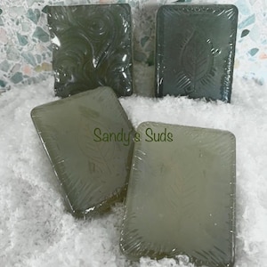 May include: Four bars of green soap with different designs. The soap is wrapped in clear plastic and the text "Sandy's Suds" is visible on one of the bars.