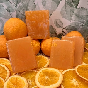 May include: Four orange-scented soap bars arranged on a bed of dried orange slices. The soap bars are a light orange color and have a subtle swirl pattern. The text "Sandy's Suds" is visible on one of the soap bars.