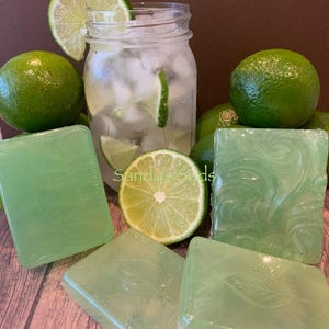 May include: A refreshing composition featuring green limes, ice, and a mason jar filled with a beverage. Several bars of green soap with leaf and swirl designs are arranged around the limes. The text "Sandy's Suds" is visible.