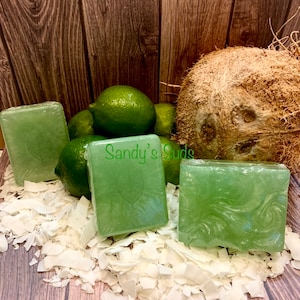 May include: Three green bars of soap with a coconut and lime scent. The soap is wrapped in clear plastic and has a swirl design. The soap is sitting on a bed of shredded coconut. The soap is labeled "Sandy's Suds".