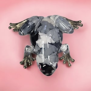 Pode incluir: Uma figura decorativa de sapo com um padrão salpicado de cinza, branco e preto. O sapo tem patas detalhadas de cor prateada e está sobre um fundo rosa suave. Um item decorativo único.