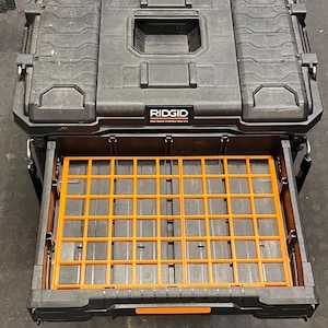 May include: Black Ridgid Pro Gear System 2.0 tool box with an orange grid insert in the open drawer. The tool box has a textured surface and a handle on top. The Ridgid logo is visible on the front.