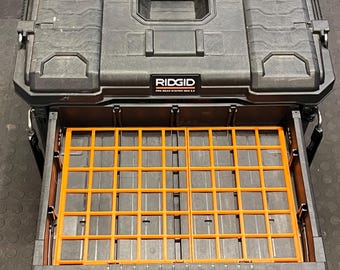 Könnte beinhalten: Schwarze Ridgid Pro Gear System 2.0 Werkzeugkiste mit einem orangefarbenen Gittereinsatz in der offenen Schublade. Die Werkzeugkiste hat eine strukturierte Oberfläche und einen Griff oben. Das Ridgid-Logo ist auf der Vorderseite sichtbar.