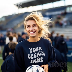 Puede incluir: Una mujer lleva una sudadera azul marino con el texto "YOUR DESIGN HERE" en blanco. Está sosteniendo un balón de fútbol blanco y negro y sonriendo.