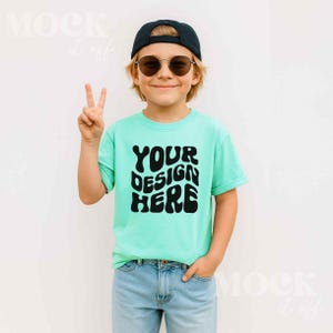 May include: A child wearing a light green t-shirt with the text "YOUR DESIGN HERE" in black, paired with blue jeans, a black cap, and sunglasses. The child is giving a peace sign.