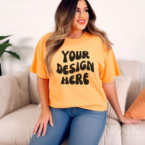 May include: Orange t-shirt with the text "YOUR DESIGN HERE" in black, displayed on a person. The shirt is paired with blue jeans. The person is seated on a beige sofa, with an orange pillow.