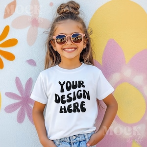 Könnte beinhalten: Ein weißes T-Shirt mit kurzen Ärmeln und einem schwarzen Aufdruck, der "YOUR DESIGN HERE" in einer Retro-Schriftart zeigt. Das Shirt wird von einer jungen Person mit braunen Haaren und Sonnenbrille getragen.