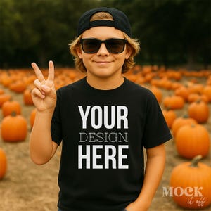 Op de afbeelding: Zwart t-shirt met de witte tekst "YOUR DESIGN HERE", gedragen door een persoon met een zwarte pet en zonnebril. De achtergrond toont een pompoenveld met oranje pompoenen.
