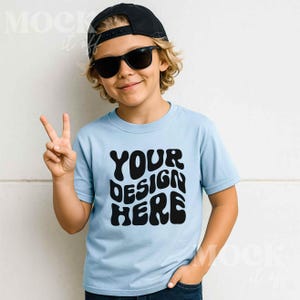 Puede incluir: Una camiseta azul claro con el texto "YOUR DESIGN HERE" en negro. La persona que la lleva puesta lleva una gorra negra y gafas de sol. La persona sonríe y hace el signo de la paz.