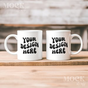 May include: Two white ceramic mugs with black text that reads "Your Design Here" on a wooden surface.