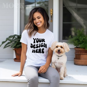 May include: White t-shirt with the text "YOUR DESIGN HERE" in black, paired with gray pants. A small, light-colored dog with a black collar sits beside the person. The scene is set on a white porch.