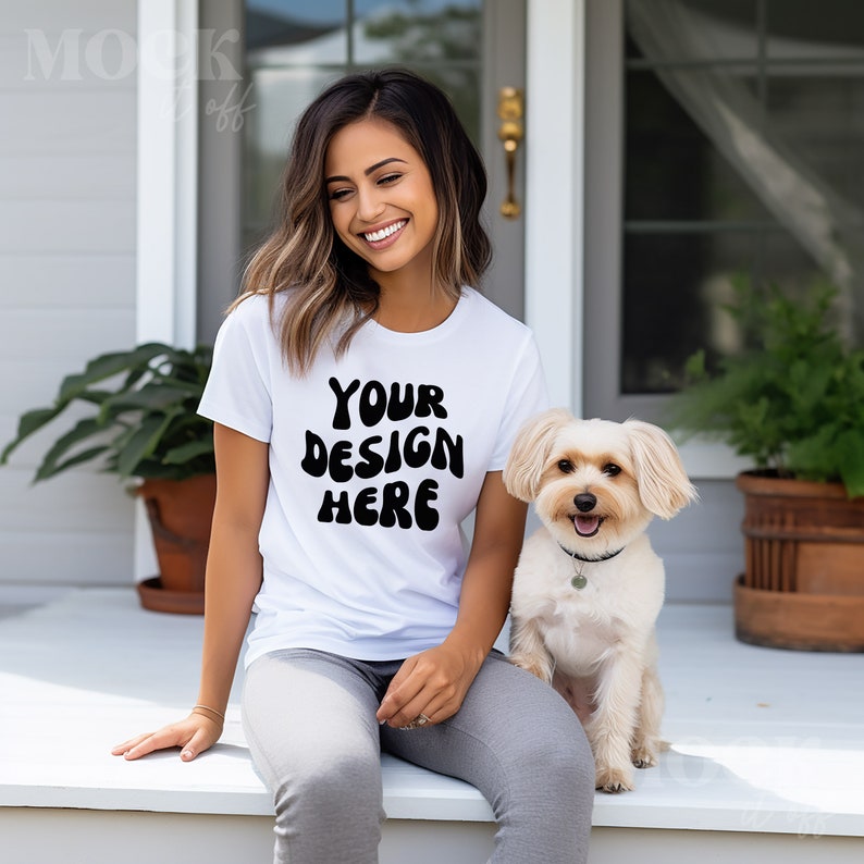 May include: White t-shirt with the text "YOUR DESIGN HERE" in black, paired with gray pants. A small, light-colored dog with a black collar sits beside the person. The scene is set on a white porch.